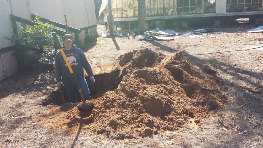Crew member digging a septic tank lid open by hand in a sandy South Carolina yard